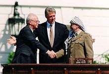 Un hombre con un traje oscuro de la izquierda da la mano de un hombre en el tocado tradicional árabe a la derecha. Otro hombre (Bill Clinton) está de pie con los brazos abiertos en el centro detrás de ellos.