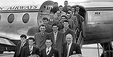 Una fotografía en blanco y negro de varias personas en trajes y abrigos en las gradas de un avión.