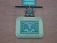 Una tableta de piedra, inscrita con la imagen de un campo de fútbol y varios nombres. Está rodeado por una frontera de piedra en forma de un estadio de fútbol. Por encima de la tableta es una talla de madera de dos hombres que sostienen una gran corona.