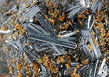 Una vista de cerca de una roca en costra con grupos de vidrioso, lustroso, Hutchinsonita plateado-azul, en racimos apretados de cristales en forma de aguja ligeramente alineados, entre pequeños grupos de cristales de color naranja-marrón diminutos