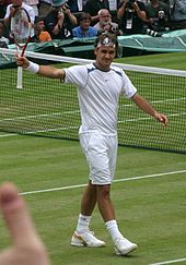 Un hombre de cabello oscuro está agitando a la multitud con su raqueta de tenis en la mano derecha, y él está usando toda la ropa blanca