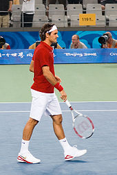 Un hombre de pelo oscuro se encuentra en una camisa roja con pantalones cortos y zapatos blancos y pañuelo, que esta persona lleve su raqueta de tenis en la mano derecha apuntando hacia el suelo