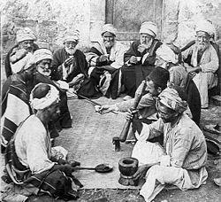 Una vieja foto de una docena de hombres viejos y de mediana edad que se sientan en el suelo alrededor de una alfombra. Un hombre frente se sienta junto a un mortero y tiene un bate, listo para la molienda. Un hombre frente a él tiene una cuchara larga.