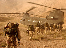 Una línea de soldados con equipo a la espalda para caminar hacia un helicóptero de transporte en terreno desértico