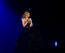 Kylie wears a feather dress with a gold corset on, holding a microphone looking towards her audience. She is also wearing a hat standing in front of a dark blue and black background (2011).