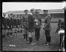 Una foto en blanco y negro de un campo de rugby en el que tres hombres vestidos con uniforme militar, uno de los cuales es el rey Jorge, presentan un trofeo de plata a un jugador de rugby vestido con kit negro. Detrás de una línea son el resto del equipo.