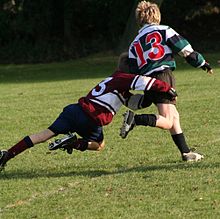 Un niño que corre lejos de cámara en camiseta de rugby hooped verde y negro está en proceso de ser abordado en torno a las caderas y las piernas por un oponente.