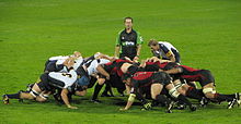 Dos formaciones opuestas de ocho hombres, en blanco y negro a la izquierda, rojo y negro a la derecha, empujar contra otros en una posición de cuclillas; detrás de ellos se encuentra otro jugador y el árbitro.
