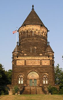 Victorian Gothic monument in brown stone. The first floor is square and the second is cylindrical with a conical roof.