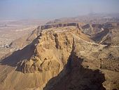 Ruinas en la cima plana de una montaña de color arena, rodeado de desierto. Otras montañas son visibles en el fondo.