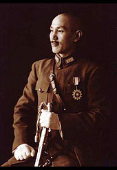 Un hombre chino en uniforme militar, sonriendo y mirando hacia la izquierda. Él sostiene una espada en su mano izquierda y tiene una medalla en forma de un sol en el pecho.
