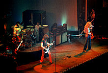 Una fotografía en colores de los cuatro miembros de Led Zeppelin ejecución en el escenario, con algunas otras figuras visibles en el fondo