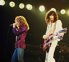 Una fotografía en colores de Robert Plant con micrófono y Jimmy Page con una guitarra de doble cuello actuando en el escenario.