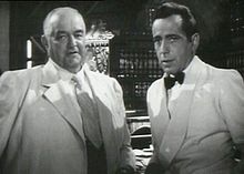 Blanco y negro película de captura de pantalla de dos hombres, ambos vistiendo trajes. El hombre de la izquierda es mayor y es casi calvo; el hombre de la derecha tiene el pelo negro. En el fondo varias botellas de alcohol pueden ser vistos.