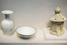 Tres piezas de porcelana blanca. From left to right, a vase with a grooved, fat body, a short, thin stem, and small opening with a large flat rim extending from it, a simple bowl with a thin brown trim around the outside rim, and a container in the shape of a granary, with several viable support posts, a tiled roof, and a staircase leading to a door, all built into the piece.