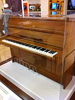 Una imagen de un piano vertical de tamaño mediano de color marrón en una caja de cristal. Las teclas del piano están expuestos.