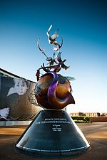 Una fotografía en colores de un monumento grande de metal con una base cónica que soporta un globo que se envuelve en instrumentos musicales contorsionadas. En el fondo es un cielo azul.