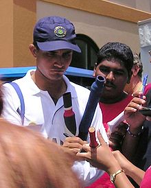 Un hombre de mediana edad que firma en palos de cricket. Lleva una camiseta blanca y una gorra azul marino. Un número de personas no son visibles, que le rodean.