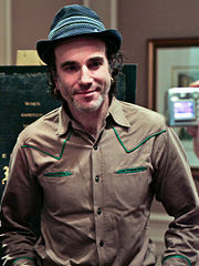 Un hombre sonriente con un sombrero gris con ribetes encima de la banda, y una camisa de estilo occidental bronceado, se encuentra en una oficina, posando para la cámara.