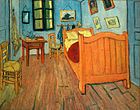 Una habitación estrecha con suelo de madera, paredes verdes, una cama grande a la derecha, a 2 sillas de paja a la izquierda, y una pequeña mesa, un espejo y una ventana cerrada en la pared trasera. Se cierne sobre la cama varias pequeñas imágenes