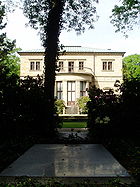 Une dalle grise, au niveau du sol encadrée par des buissons et dans l'ombre de l'arbre. Dans le fond d'une fontaine et une grande maison de deux étages avec un balcon.