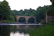 Un pont en pierre à deux arches à travers une rivière, vue le long de la rivière, les deux extrémités cachés par les arbres. Un déversoir se trouve en face du pont, à l'extrémité droite duquel se trouve un immeuble à deux étages.