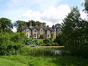 Maison de campagne, partiellement caché par la verdure, vu à travers un étang. La façade observable comprend cinq pignons, avec une tourelle entre les quatre pignons sur la gauche et la droite pignon.