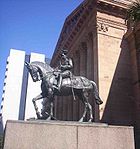 Statue équestre dans le métal gris foncé de George V en robe uniforme militaire sur un socle de granit rouge extérieur d'un bâtiment classique de grès rouge