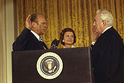 Un homme en costume, sa main droite en l'air, se tient près de sa femme et parle à un autre homme dans les robes d'un juge. Le groupe se tient devant un rideau, derrière un podium portant le sceau du président des États-Unis.