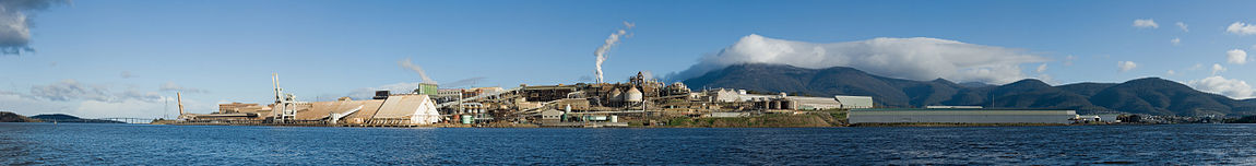 Un panorama avec une grande installation industrielle sur un côté de la mer, en face de montagnes.