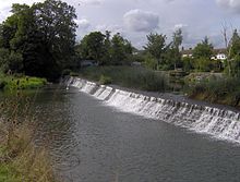 Un barrage avec de l'eau qui coule de droite à gauche, entouré d'arbres et de la végétation.