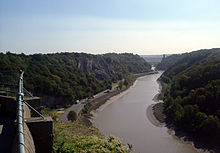 Rivière se écoulant à travers une vallée encaissée. Dans la distance est un pont suspendu soutenu par des pylônes. Dans le premier plan à gauche est une main courante.