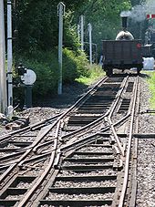 Une voie ferrée avec l'éloignement où un train à vapeur se; la piste a trois rails, au milieu de laquelle est décalé vers la droite au premier plan, mais passe à gauche au milieu à un pointwork complexe où trois autres rails joignent de la gauche