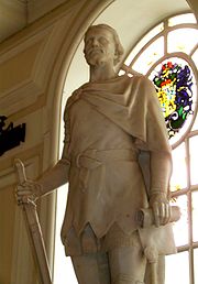 La statue d'un homme dans une cape courte tunique et serré à son épaule droite, sculptée en pierre blanche. Le chiffre, fixé à l'intérieur avec le dos à une fenêtre cintrée, tient une épée de bas-pointu dans sa main droite et un parchemin dans sa gauche.