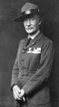 Photographie en noir et blanc d'un homme en uniforme militaire, avec ses médailles épinglées sur le côté gauche de sa veste. Il porte un chapeau à large bord et tenant un bâton de marche dans les deux mains.