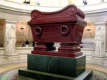 Photo of a large, shiny burgundy cuboid-shaped vessel raised on a dark green plinth. There are two female statues in the background either side of the vessel.
