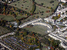 Photographie aérienne de terrasse semi-circulaire de bâtiments en pierre avec grande étendue d'herbe en face et à gauche. Montre également terrasses de bâtiments environnants.