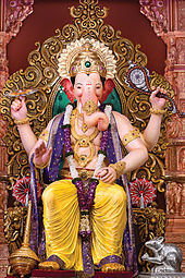 A sitting elephant-headed four-armed man statue, wearing gold ornaments, flower garlands and an orange dhoti.