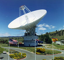 White satellite dish pointed upwards with United States, Australian, and Spanish flags in foreground