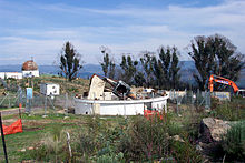 White circular building missing roof, with large metal debris in center, surrounded by chain-link fence