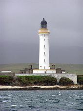 Un grand phare blanc avec une bande brune autour du parapet et la lanterne de couleur sombre se asseoir sur une côte rocheuse. Un mur blanc obscurcit l'étage inférieur de bâtiments de pierre grise réunis autour de sa base.