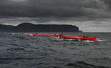 Un long tube rouge se trouve dans l'eau sous un ciel sombre, couvertes de nuages avec des collines noires dans la distance.