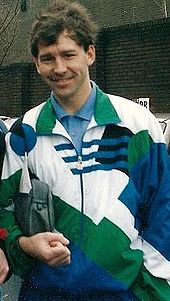 Un homme souriant avec des cheveux noirs portant un blanc, vert et bleu haut de survêtement sur une chemise bleue. Il tient une trousse de toilette sous son bras droit.