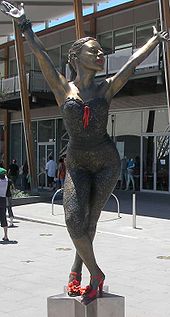 Une statue de bronze de Kylie, sur un piédestal en forme d'étoile, la représente dans une pose danse. Ses jambes sont croisées et elle se penche à la taille, avec les deux bras tendus au-dessus de sa tête. La statue se dresse sur une place publique en face d'un bâtiment moderne de verre et plusieurs personnes ne marche.