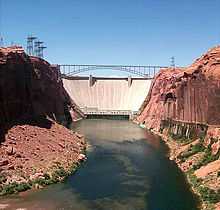 Un barrage presque blanc se étend à la roche de couleur rouge de chaque côté. An arching steel bridge crosses in front of the dam.