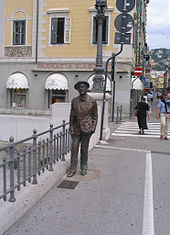 Statue de bronze de Joyce debout sur un trottoir, à côté d'un garde-corps. Derrière la statue est une scène de rue avec les piétons et les magasins.