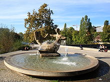 Fontaine circulaire entouré de trottoir. Le centre de la fontaine est une sculpture d'une paire de figures humaines abstraites.