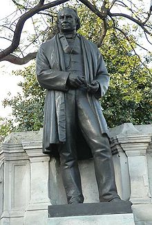 Une sculpture en bronze métallique d'un homme du XIXe siècle portant une longue veste ou un manteau, un pantalon, gilet, avec les outils de dessinateur dans ses mains