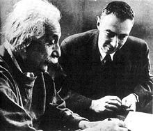 Einstein écrit à un bureau. Oppenheimer est assis à côté de lui, regardant.