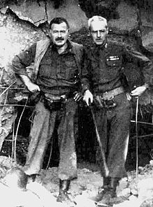 un homme aux cheveux sombres avec la moustache vêtu de bottes de l'armée, niché dans un pantalon boueux, chemise et gilet, avec un homme aux cheveux lumière vêtus de vêtements de l'armée debout en face de gravats